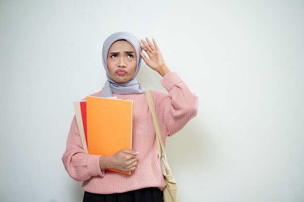 Glimlachende Aziatische moslim vrouwelijke student in roze trui met medium tas met boek en denken