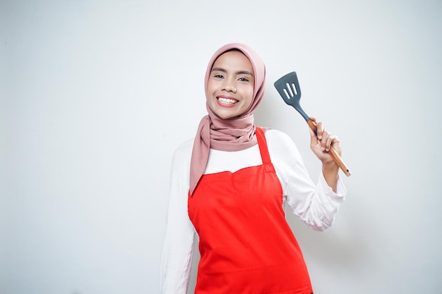Glimlachende Aziatische moslimvrouw die de rode spatel van de schortholding draagt Kookgerei Kokend concept