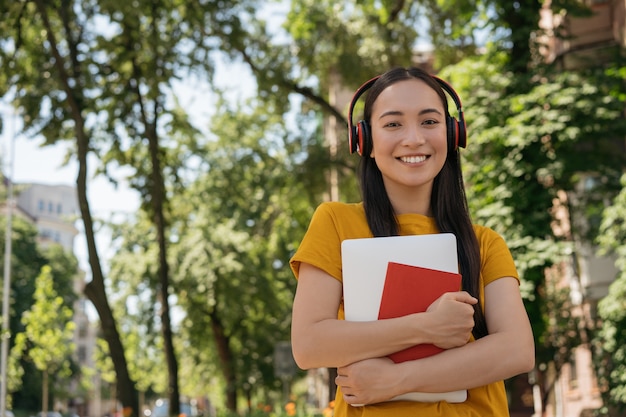 Glimlachende Aziatische student die hoofdtelefoons draagt die naar universiteit terug naar schoolconcept lopen