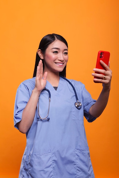 Glimlachende aziatische verzorger in telehealth videocall op afstand met patiënt terwijl ze online consultatie hebben op oranje achtergrond. Kliniekverpleegster die een stethoscoop draagt die bij teleconferentie videochatgesprek zwaait.