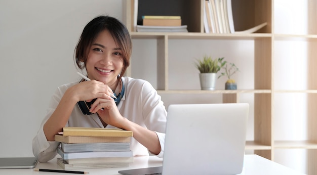 Glimlachende Aziatische vrouw die aan het bureau met boeken en laptop computer werkt.