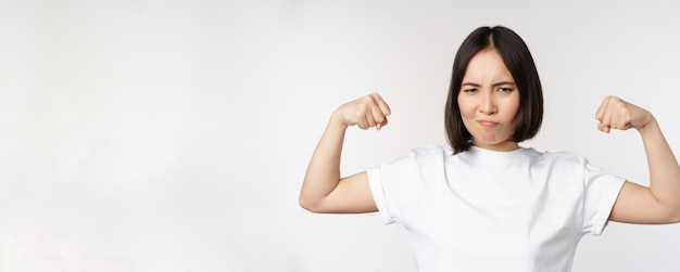 Glimlachende aziatische vrouw die buigende bicepsspieren toont, sterk armengebaar in witte t-shirt over...