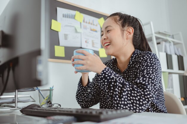 Glimlachende aziatische vrouw die een polka dot-shirt draagt met een kopje koffie of thee in de buurt van de computer op de werkplek