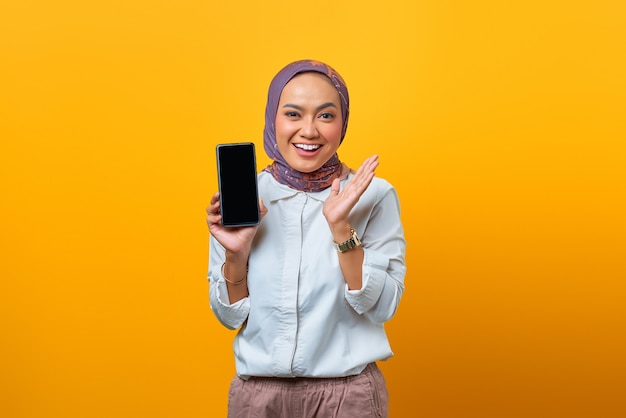 Glimlachende aziatische vrouw die het lege smartphonescherm over gele achtergrond toont