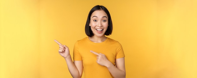 Glimlachende aziatische vrouw die met de vingers naar links wijst en advertenties toont op lege kopieerruimte die over een gele achtergrond staat