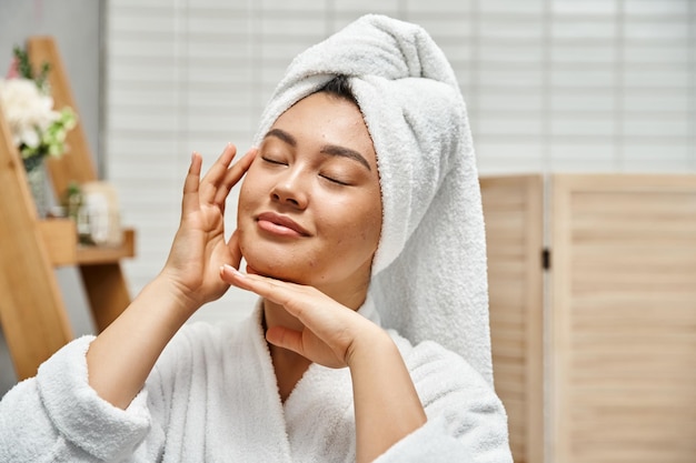 glimlachende Aziatische vrouw met acne en witte handdoek op het hoofd met gesloten ogen in de badkamer