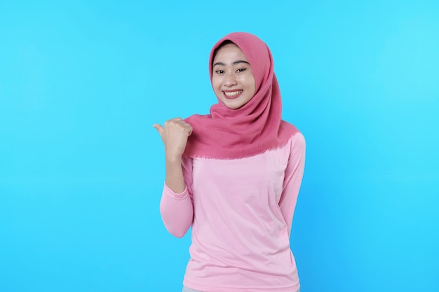 Foto glimlachende aziatische vrouw met haar duim wijzend geïsoleerd op een lichtblauwe achtergrond die hijab draagt met roze t-shirt