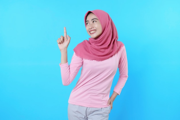 Glimlachende aziatische vrouw met haar vinger wijzend geïsoleerd op lichtblauwe achtergrond die hijab draagt met roze t-shirt