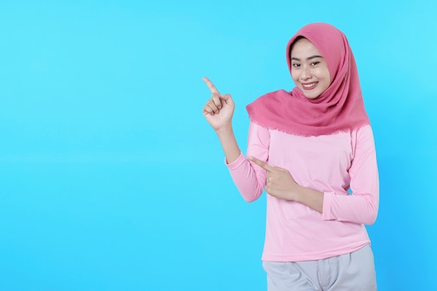 Glimlachende aziatische vrouw met haar vinger wijzend geïsoleerd op lichtblauwe achtergrond die hijab draagt met roze t-shirt