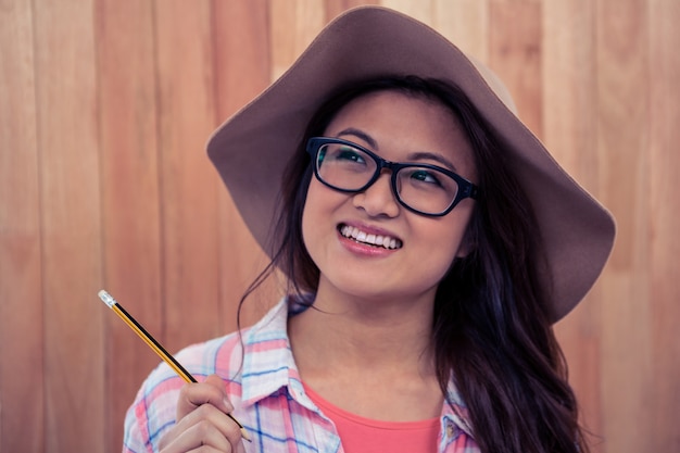 Glimlachende Aziatische vrouw met het potlood van de hoedenholding tegen houten muur