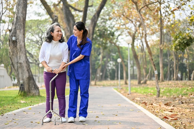 Glimlachende Aziatische vrouwelijke verzorger die een oude dame bijstaat om met een wandelstok te lopen
