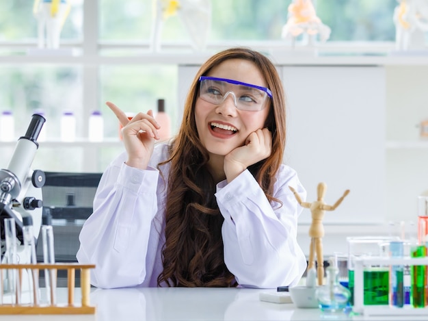 Glimlachende Aziatische vrouwelijke wetenschapper in glazen zittend aan tafel met microscoop en kolven en wijzende vinger in laboratorium tonen.
