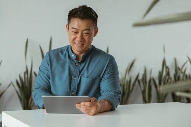 Glimlachende Aziatische zakenman die aan digitale tablet werkt terwijl hij in een gezellig kantoor zit