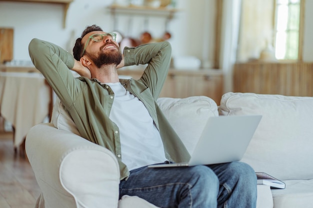 Glimlachende baarde man met een stijlvolle groene bril met een laptop die zich uitstrekt en een pauze neemt