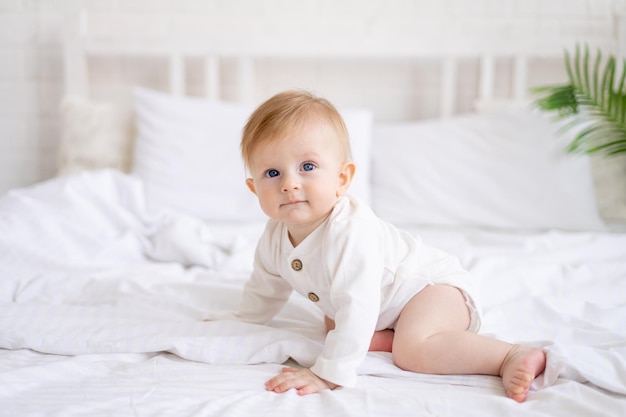 Glimlachende baby 6 maanden oude blonde jongen kruipt op een wit bed in een lichte slaapkamer na het slapen in de ochtend in een katoenen bodysuit het concept van kinderartikelen