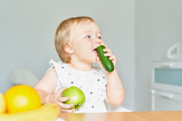 Glimlachende baby die komkommer bijt en groene appel vasthoudt terwijl hij op de kinderstoel zit en wegkijkt