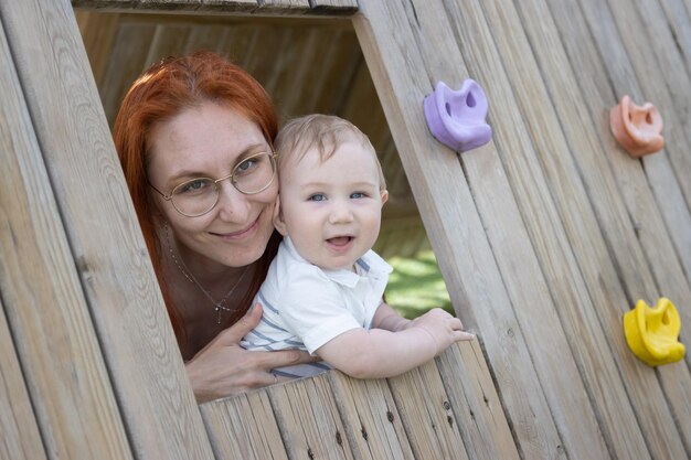 Foto glimlachende baby en zijn moeder zitten in een houten huis op de speeltuin en kijken in de camera