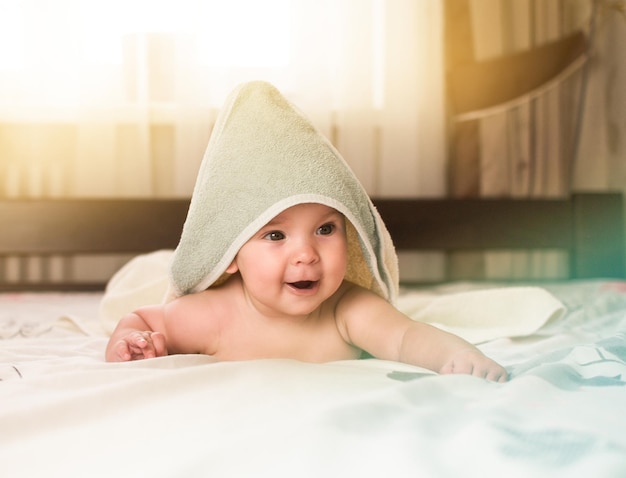 Glimlachende baby na het nemen van een douche of bad ligt op het bed met een handdoek op zijn hoofd en lichaam
