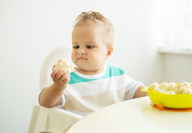 Glimlachende babyjongen zittend in een stoel Childs bloemkool eten op witte keuken.