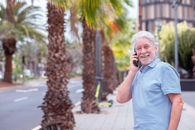 Glimlachende bebaarde blanke senior man met behulp van mobiele telefoon die buiten in de straat staat Palmbomen en gebouwen op de achtergrond