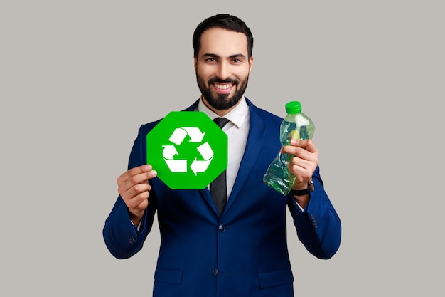 Glimlachende bebaarde man die de hand uitsteekt met een plastic fles met een bord voor recycling, het sorteren van afval ter bescherming van de natuur, het dragen van een officieel pak. Indoor studio opname geïsoleerd op een grijze achtergrond.