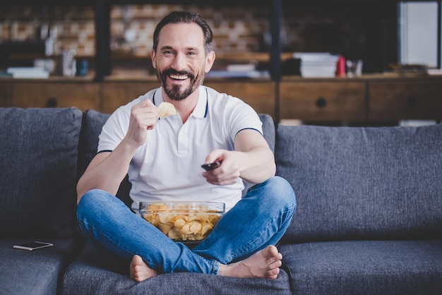 Glimlachende bebaarde man zittend op de bank tv kijken en thuis chips eten