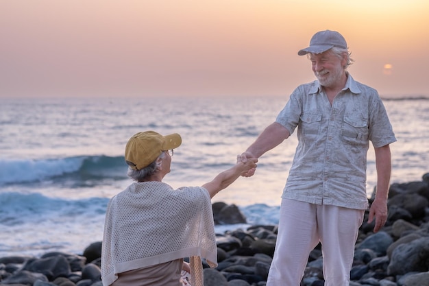 Glimlachende bejaarde man helpt zijn vrouw om op te staan van het strand Glimlachend oud senior paar buitenshuis genietend van hun vakantie samen