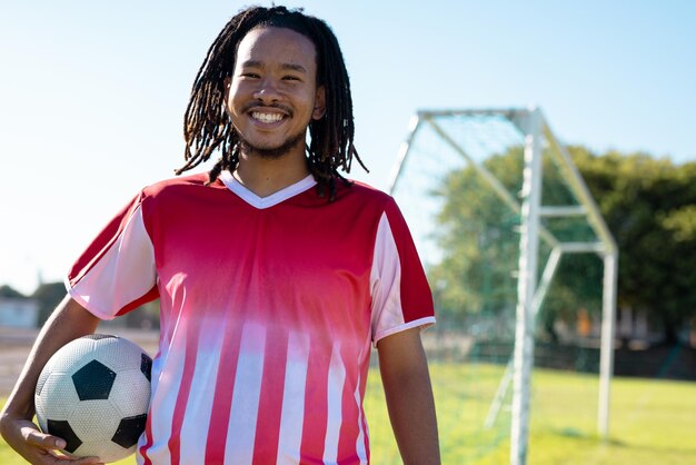 Glimlachende biraciale mannelijke speler met dreadlocks die rode trui dragen met bal die tegen heldere hemel staat