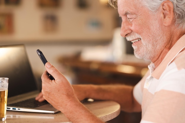 Glimlachende blanke bebaarde senior man zit aan pub tafel met een glas bier werken op laptop met behulp van telefoon verslaafde ouderen genieten van tech en sociaal