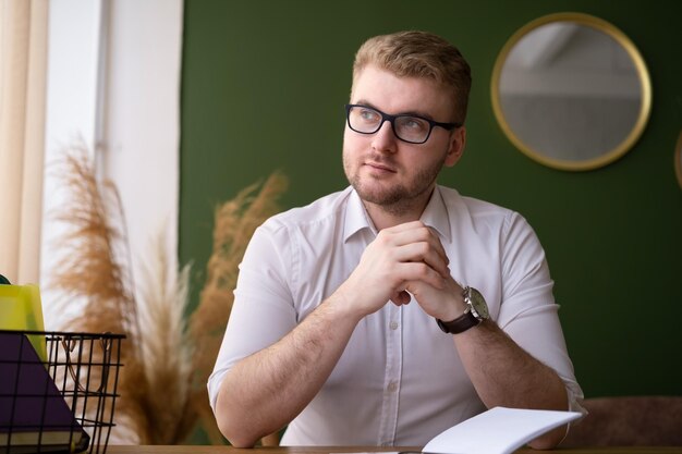 Glimlachende blanke blonde volwassen man aan het werk aan het bureau binnenshuis in een stijlvol, modern huisinterieur Ernstige doordachte zakenman op het werk