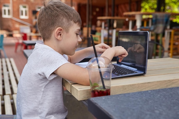 Glimlachende blanke jongen zittend aan tafel op de veranda van café studeren met laptop