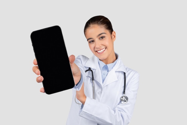 Glimlachende blanke millenniale vrouwelijke dokter in witte jas toont smartphone met leeg scherm.