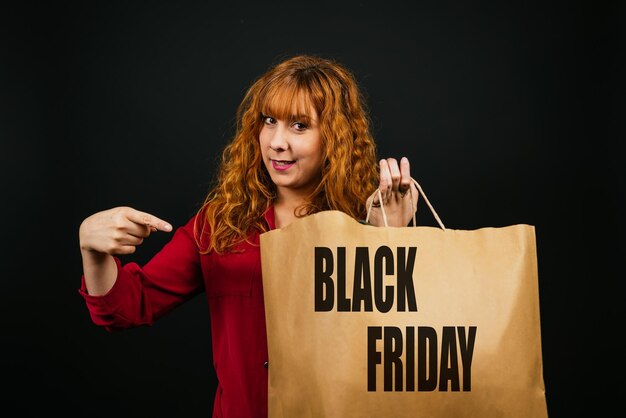 Foto glimlachende blanke vrouw die met haar vinger wijst naar een boodschappentas op een zwarte achtergrond black friday