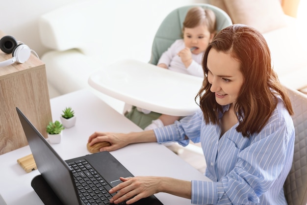 Glimlachende blanke vrouw in blauw shirt werkt op laptop, kind zit naast haar, moeder met zwangerschapsverlof