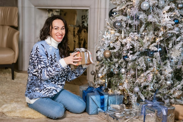 Glimlachende blanke vrouw opent doos met kerstcadeau in gebreide trui en spijkerbroek terwijl ze zit...