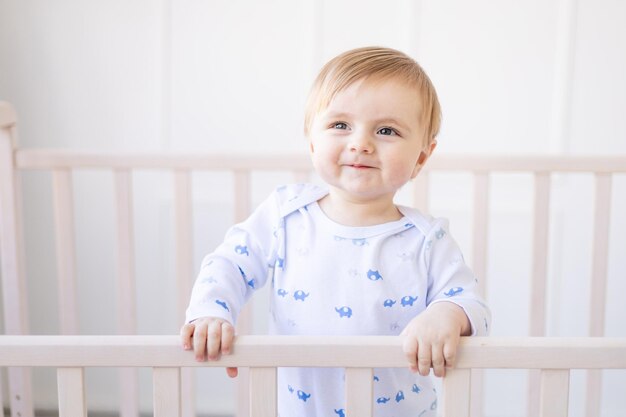 Glimlachende blonde babyjongen in een wieg thuis met blauw en wit katoenen beddengoed staat aan de kant en lacht in de ochtend het concept van kinderartikelen en accessoires