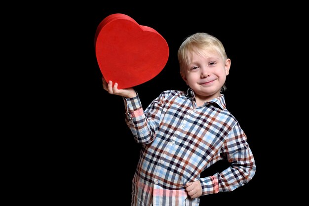 Glimlachende blonde jongen die in geruit overhemd een rode hart gevormde doos houdt.