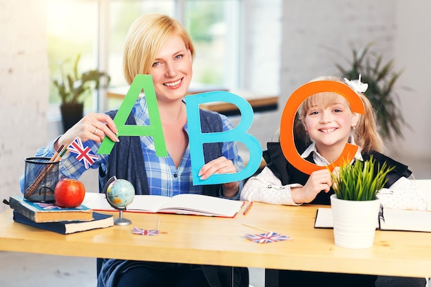 Glimlachende blonde leraar Engels speelt letters en woorden met schoolmeisjestafel met boeken in lichte klas met bloempotten