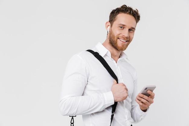 Glimlachende blonde man met behulp van mobiele telefoon en draadloze koptelefoon geïsoleerd over witte muur