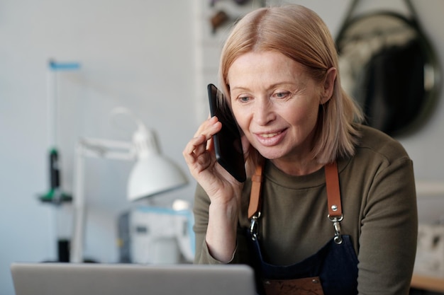 Glimlachende blonde rijpe vrouw die met smartphone door oor laptop het scherm bekijken