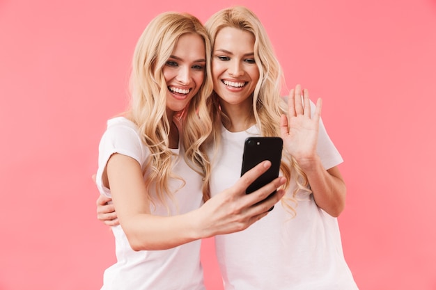 Glimlachende blonde tweeling die in t-shirts draagt die door videocall op smartphone over roze muur praten