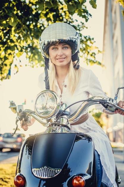 Glimlachende blonde vrouw in jeans, t-shirt en helm met haar Vespa-scooter.