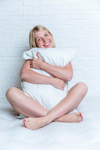 Glimlachende blonde vrouw zit in een gezellig bed knuffelen met een kussen op de achtergrond van een witte bakstenen muur Rust en luiheid Verticaal