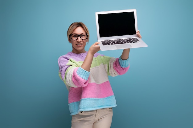 Glimlachende blonde vrouwelijke adviseur in een gestreepte trui demonstreert een mockup van een laptop op een blauwe achtergrond