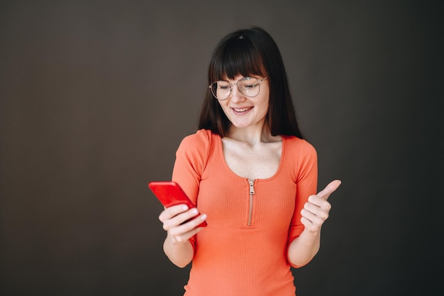 Glimlachende brunette in jurk en bril poseren geïsoleerd op zwarte achtergrond met een telefoon in haar handen