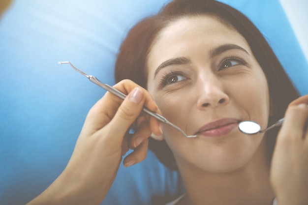 Glimlachende brunette vrouw die wordt onderzocht door een tandarts in een tandheelkundige kliniek. Handen van een arts die tandheelkunnige instrumenten bij de mond van de patiënt houden. Gezonde tanden en medicijnconcept.