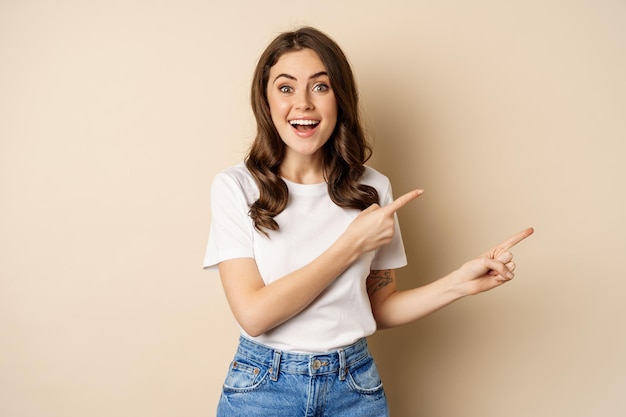 Glimlachende brunette vrouw in t-shirt, wijzende vingers naar rechts, promo-aanbieding of advertentie tonen, banner demonstreren, staande tegen een beige achtergrond