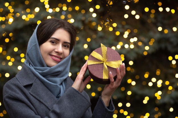 Glimlachende brunette vrouw met geschenkdoos in de buurt van de kerstboom. Ruimte voor tekst