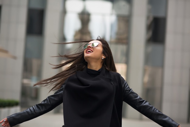 Glimlachende brunette vrouw met wapperende haren in de wind wandelen in het winkelcentrum van de stad