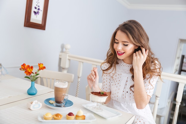 Glimlachende charmante jonge vrouw die dessert eet en latte drinkt in café